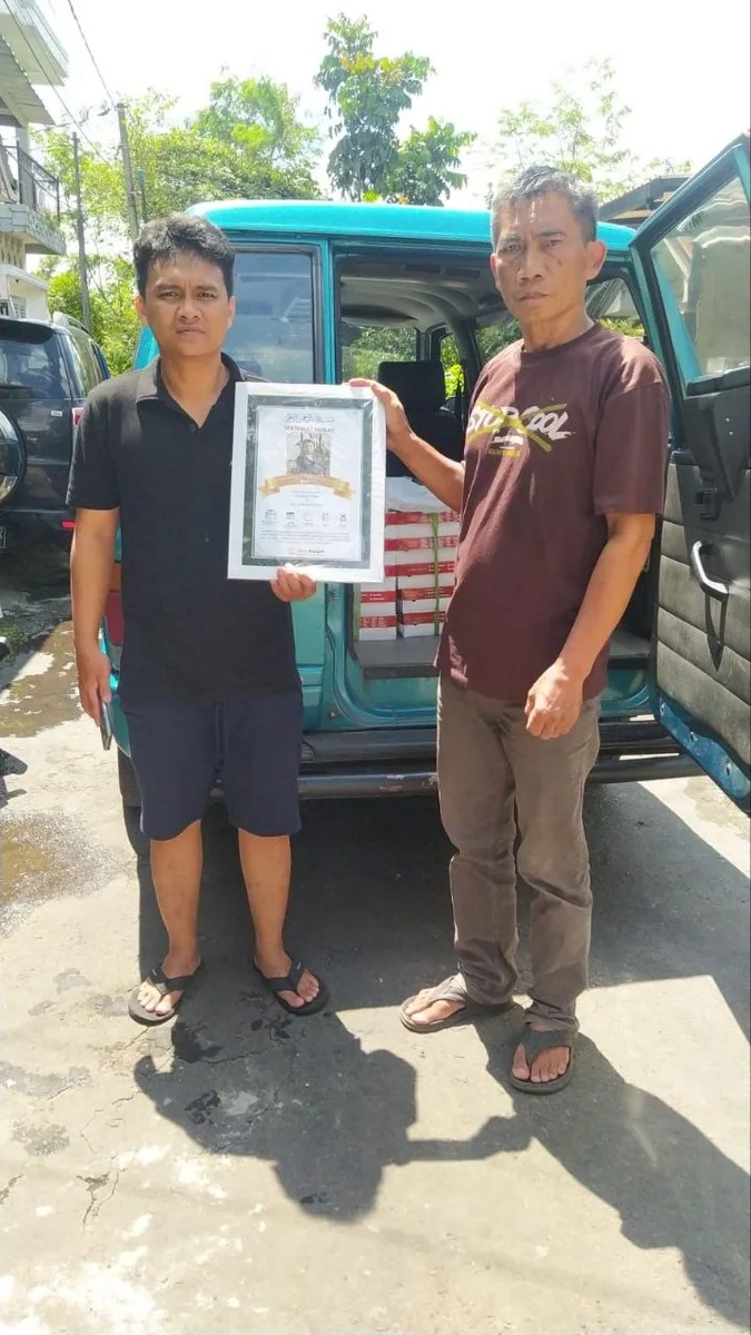 Dua pria memegang sertifikat aqiqah di depan kendaraan berisi box Quy